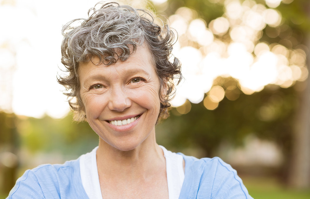 A smiling person with short curly hair stands outdoors, surrounded by blurred trees and sunlight.